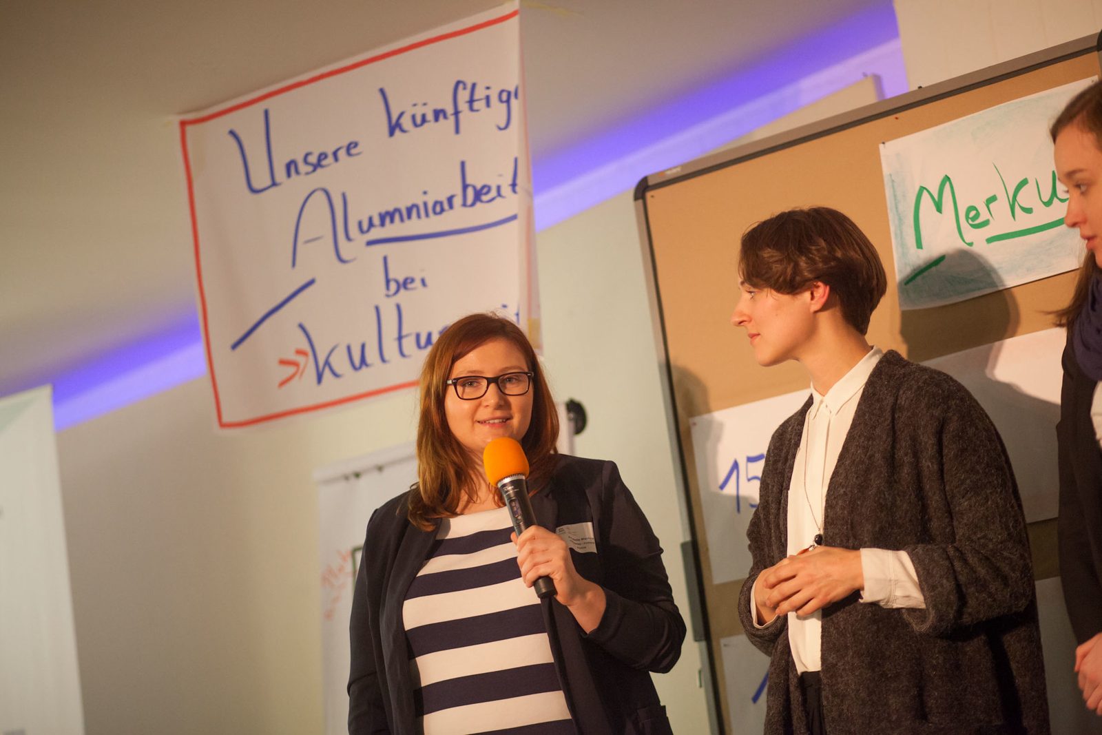 Zwei Alumni sprechen mit Mikrofon vor einem Plakat mit der Aufschrift „Unsere künftige Alumniarbeit bei kulturweit“
