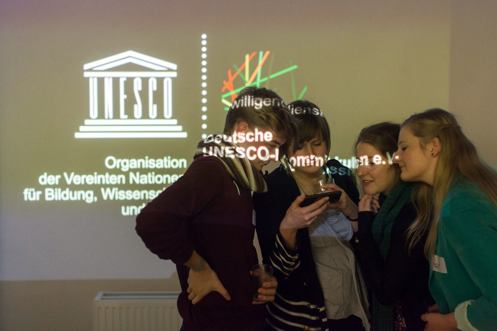 Vier Alumni schauen gemeinsam auf ein Handy, hinter ihnen sind die Logos der UNESCO und von kulturweit an die Wand projiziert