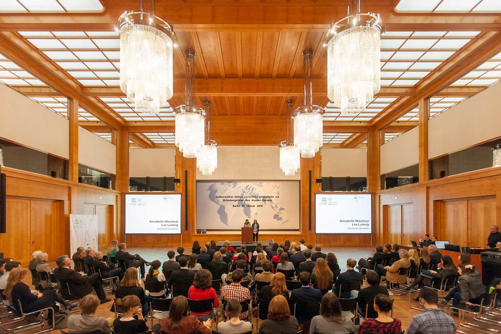 Voller Saal mit Holzvertäfelung und Kronleuchtern, vorne die Leinwand „Gründungsfeier des Alumni-Vereins, Berlin, 30. Januar 2015“