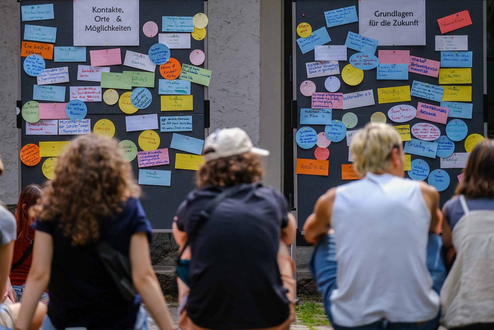 Alumni sitzen vor zwei Pinnwänden voller bunter Karten mit den Überschriften „Kontakte, Orte & Möglichkeiten“ und „Grundlagen für die Zukunft“