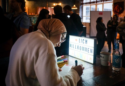 Eine Alumna schreibt an einem Tisch ihr Namensschild, daneben ein Leuchtschild mit „Herzlich Willkommen Alumni“